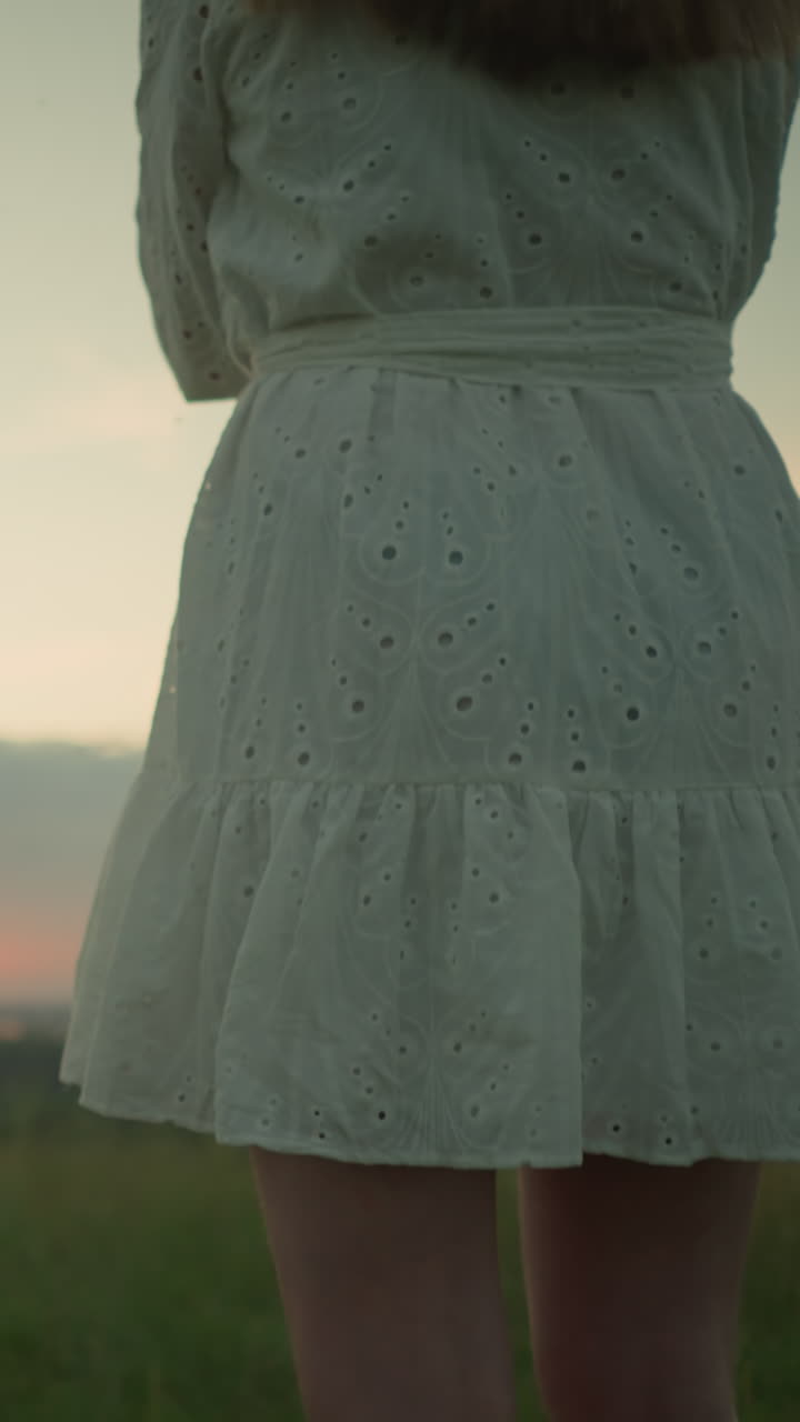 Low-angle shot on the lower body of a woman in a white dress standing alone in a serene grassy field, arms folded, as she gazes over a tranquil lake during a peaceful sunset calmness and contemplation