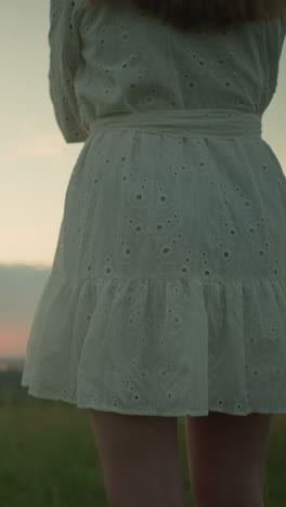 low-angle shot on the lower body of a woman in a white dress standing alone in a serene grassy field, arms folded, as she gazes over a tranquil lake during a peaceful sunset calmness and contemplation