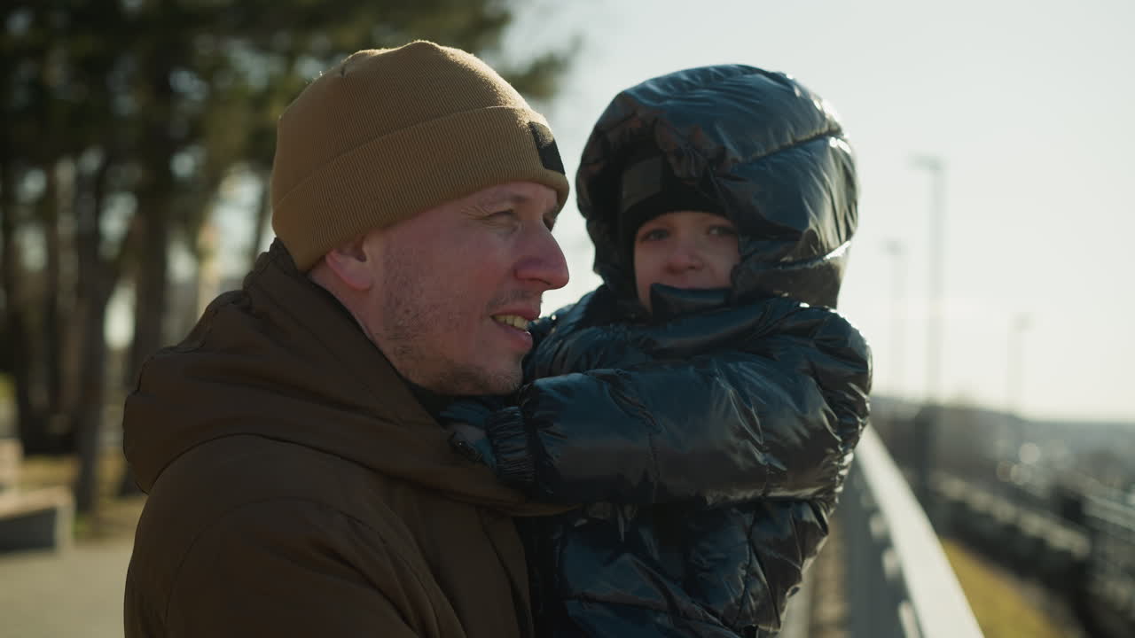 A father stands close to a metal railing, carrying son in his arms, the father gazes over a cityscape while his son looks directly at the camera, both dressed warmly in jackets and beanies