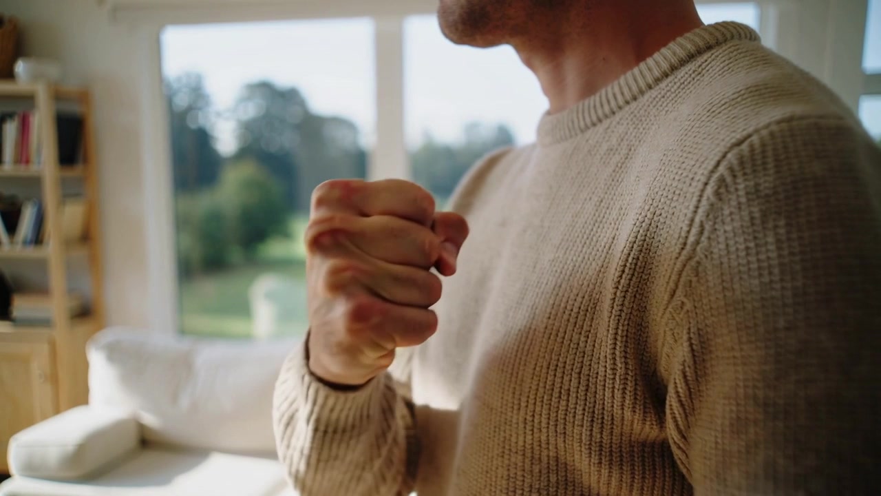 Reacting to tension, man clenching then lowering fist at home window, beige sweater, copy space