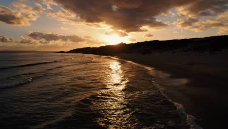 Drone-shot-of-a-golden-sunset-over-the-water-on-the-coast-of-Tarifa,-Spain