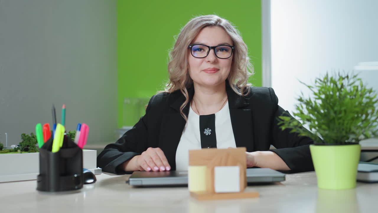 entrepreneur blond woman wearing glasses confidently closes laptop with warm smile at desk featuring building model plant pen holder notebook modern office vibe