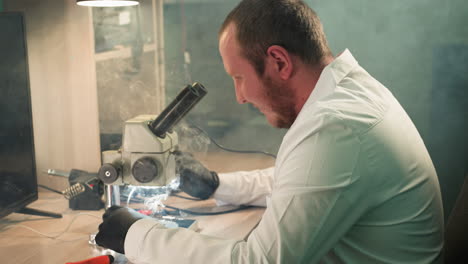 una vista de cerca de un técnico en una bata de laboratorio blanca trabajando en una placa de circuitos electrónicos bajo un microscopio, con humo del soldador llenar el lugar