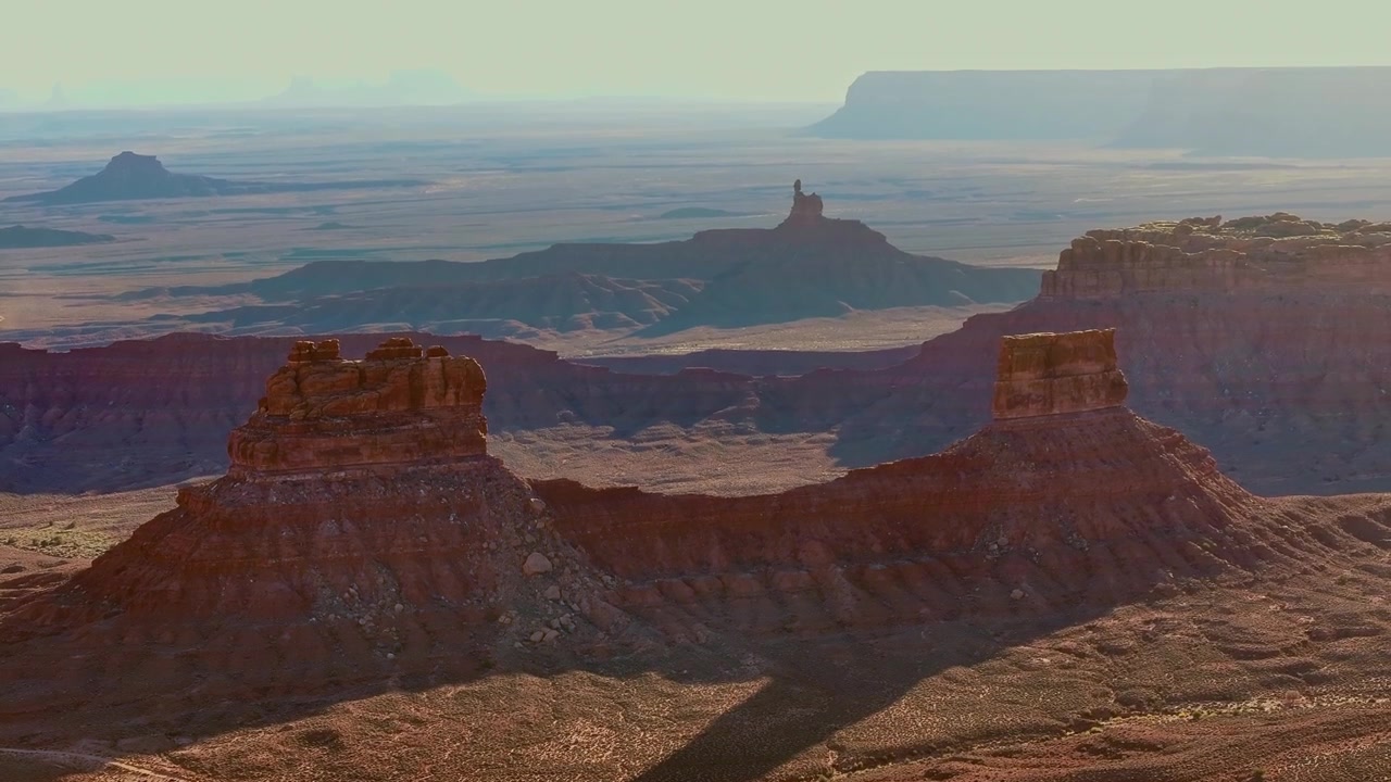 Telephoto aerial view of layered sandstone mesas and buttes at Valley of the Gods, Utah, with Rooster Butte and distant plateaus fading into the hazy horizon.