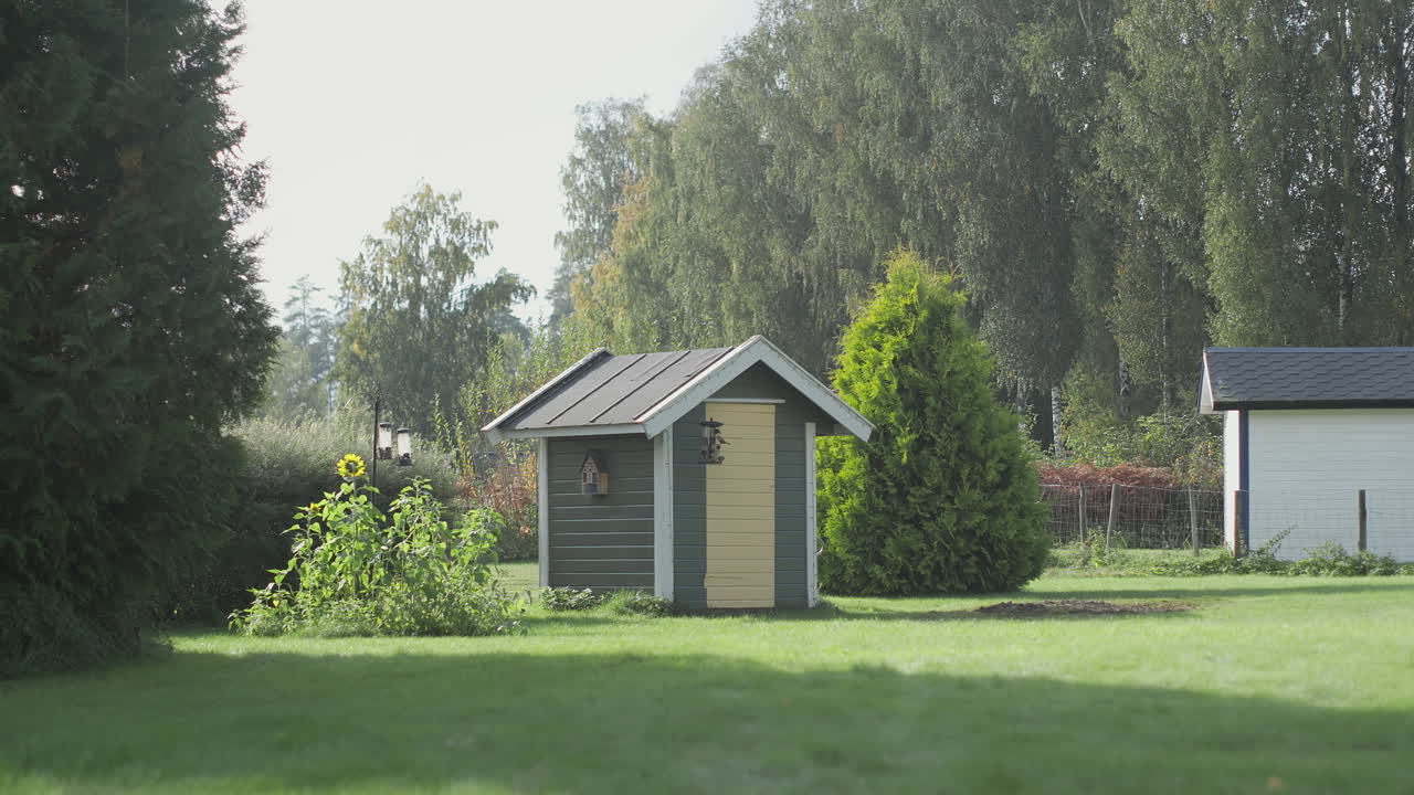 A quaint wooden shed with a pitched roof, nestled in a vibrant garden with a sunflower and tall trees nearby.