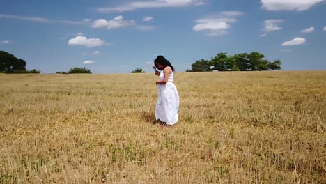 young pregnant woman in a white summer dress standing on a field holding her belly and flowers for a maternity photo shoot - drone circle close