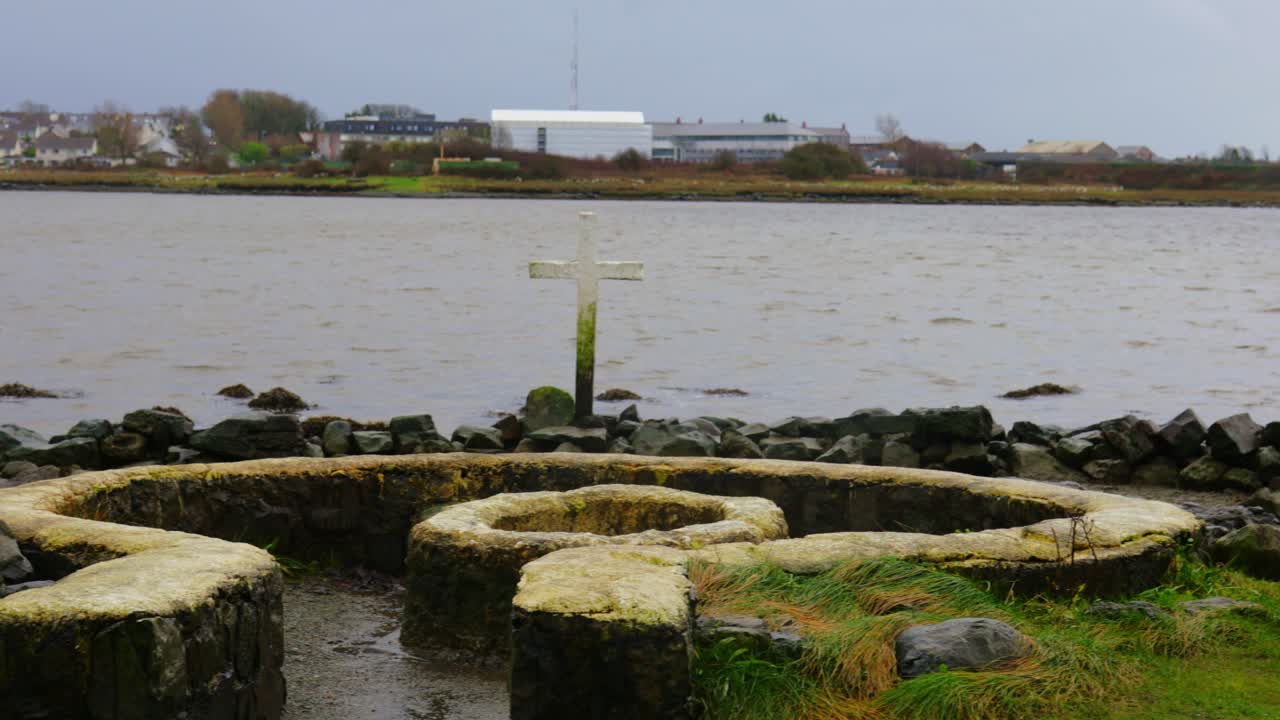 Establishing pan shot van de historische, heilige steenstructuur en het kruis van St. Augustine's Holy Well met uitzicht op Galway Bay
