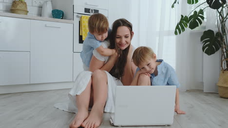 Una-Madre-Amante-De-Un-Apartamento-Moderno-Y-Dos-Hijos-Pequeños-Sentados-En-El-Suelo-De-La-Sala-De-Estar-Miran-La-Pantalla-Del-Portátil.-Los-Niños-Con-Mamá-Juegan-En-Una-Computadora-Portátil.