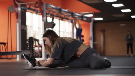 chica de cabello largo con camiseta gris y leggings hace ejercicios en alfombra en un gimnasio moderno de color naranja brillante con equipos y ventana en el fondo. estilo de vida saludable. estiramiento, inclinación del torso hacia adelante mientras estira las piernas en alfombra