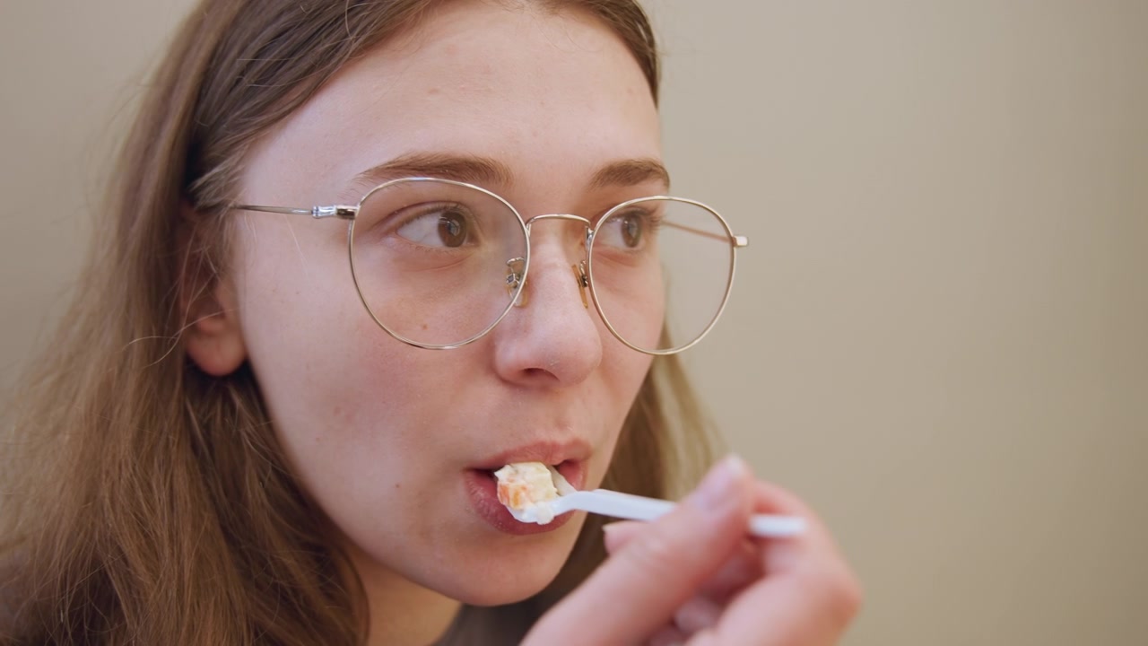 Ung kvinne smaker på en skje og grimaserer av den sure smaken i nærbilde. En brillekledd kvinne smaker på en skje innendørs, med dempet belysning og en kjøkkentest-stemning, viser ærlig ansiktsavsky og trutmunn, en naturlig følelse.