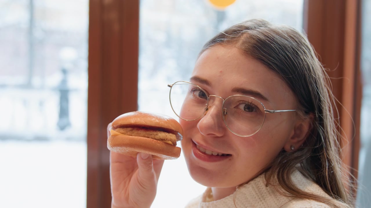 Young woman with clear glasses lifts sandwich toward mouth while smiling beside snowy window, calm indoor mood with hand movement, soft light, relaxed posture, and clear hunger anticipation