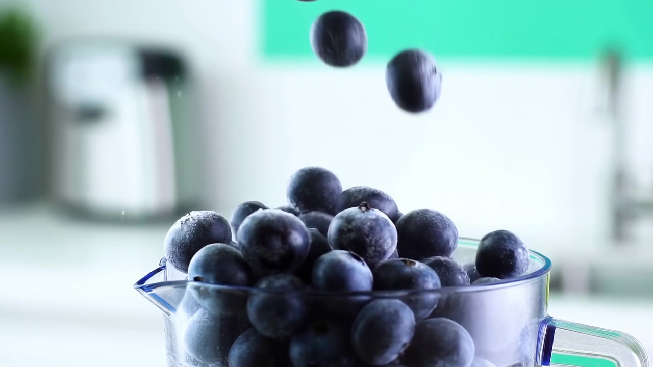 Arándanos frescos caen en una taza medidora transparente en una cocina limpia y bien iluminada, creando una escena sencilla de preparación de alimentos con un suave desenfoque de fondo.