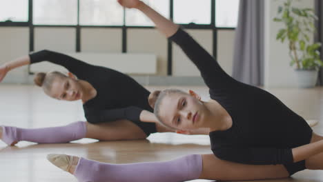 porträt von zwei jungen blonden gymnastikmädchen, die in der ballettklasse 1 dehnübungen auf dem boden machen