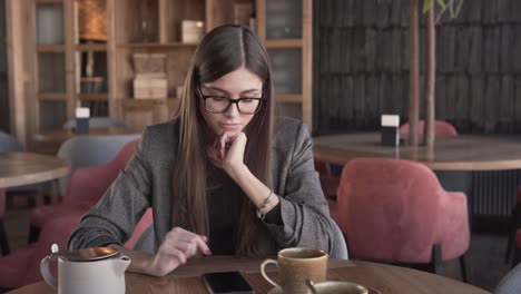 charming young woman in a glasses and smart suit is sitting in a cafe, turning the music on her phone on and dancing