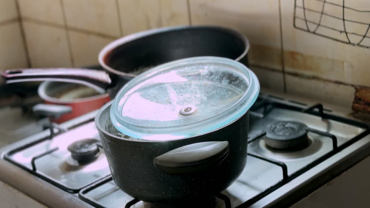 Close-up of cooking pots on a gas stove with a glass lid, saucepan, and worn tiled kitchen wall, showing everyday home cooking in an indoor kitchen setting