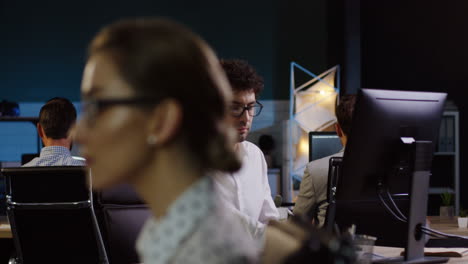 hombre de negocios con camisa blanca y anteojos trabajando tarde en la noche en la computadora rodeado de otros empleados en la oficina