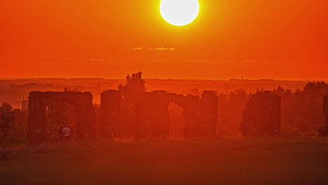 Static-shot-of-tourists-going-around-an-old-historic-building-with-the-view-of-sun-going-down-in-the-background-over-the-red-sky