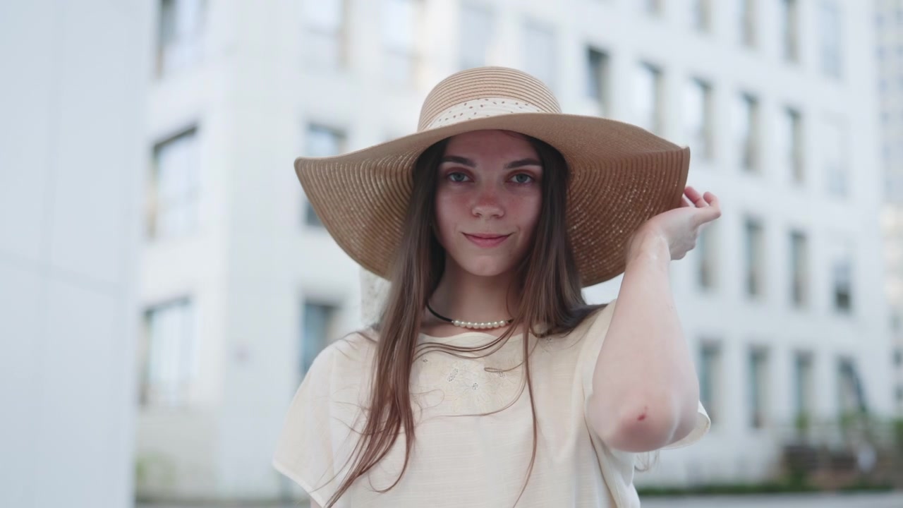 Vista laterale di una giovane donna che indossa un cappello a tesa larga e una camicia chiara, in piedi in una strada urbana con edifici sfocati sullo sfondo sotto la luce naturale del giorno.
