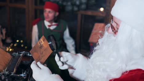 santa claus at the table looks through letters through a magnifying glass. elf is played on the background