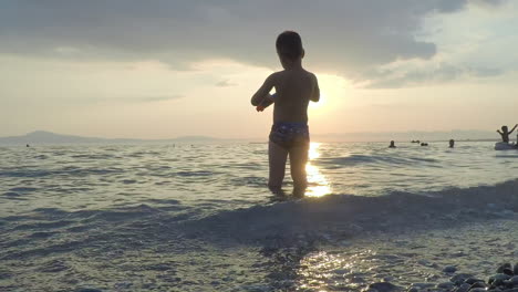 Niño-Juega-En-Las-Olas-Con-Puesta-De-Sol-En-El-Fondo-En-La-Playa-De-Kalamata,-Grecia