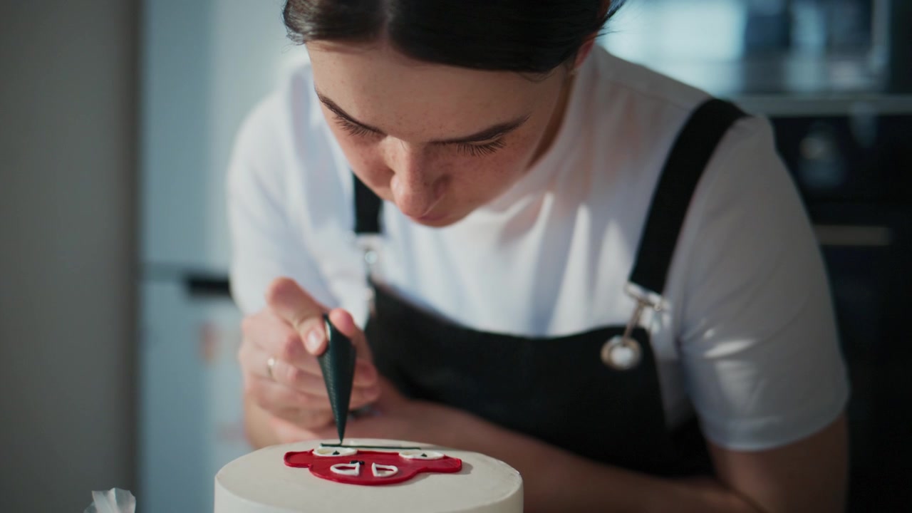 Fokusert kvinne dekorerer en kake med presisjon. Hun lener seg nært, øynene er rettet mot designet hennes. Hendene hennes beveger seg stødig, og påfører glasur med omhu. Scenen fremhever kulinarisk kunst