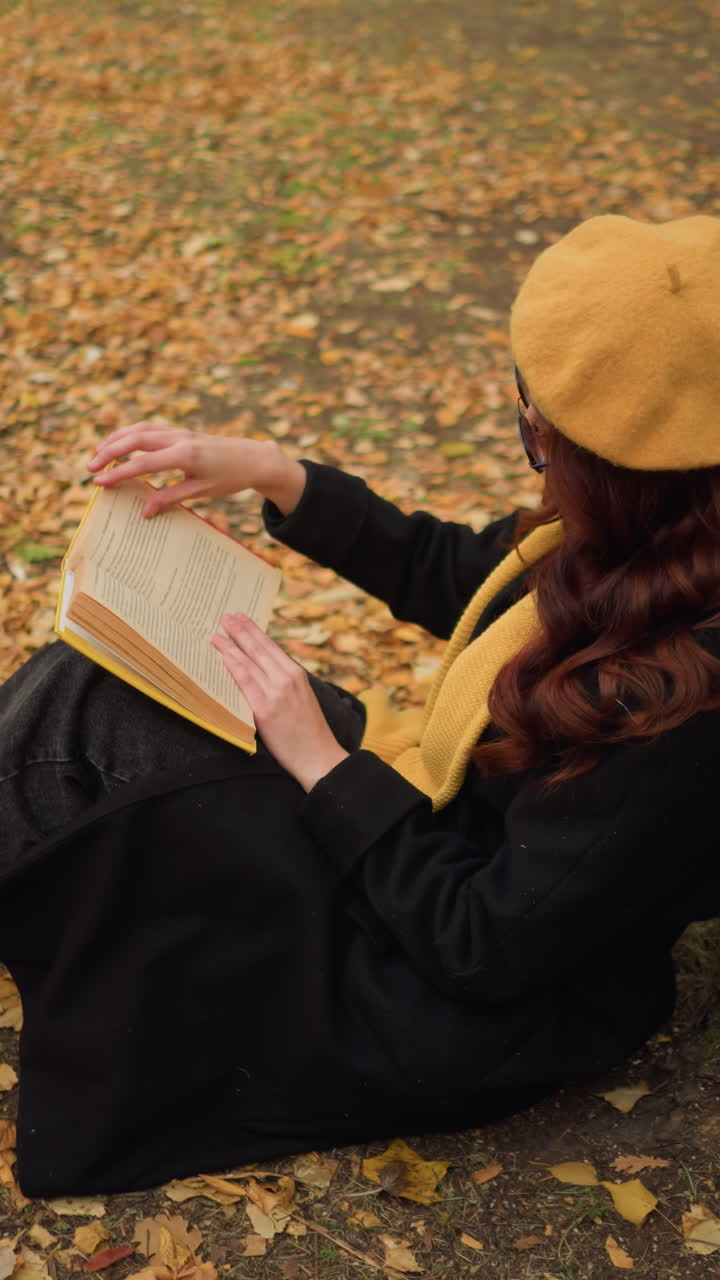 vista aérea de una mujer sentada al aire libre en el bosque de otoño apoyándose en un árbol, leyendo un libro, con una boina amarilla y un abrigo negro, desliza su mano a lo largo del borde del libro