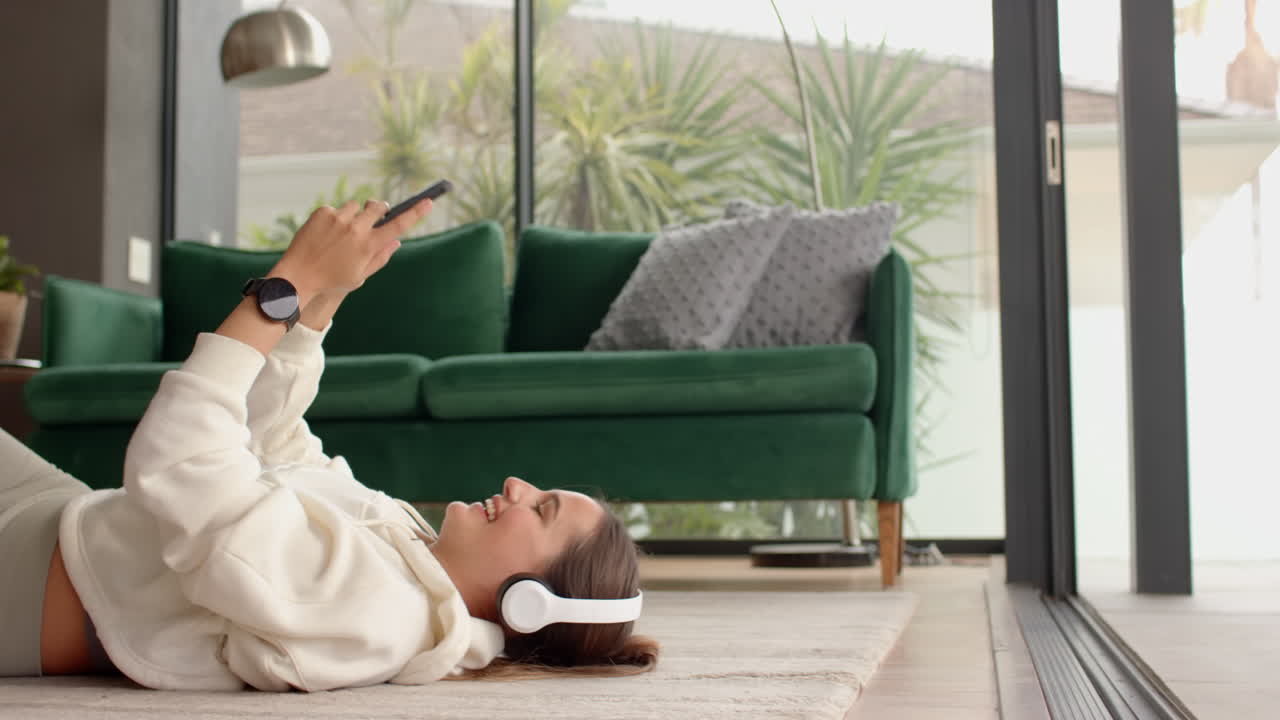 Listening to music with headphones, woman relaxing on floor holding smartphone, at home