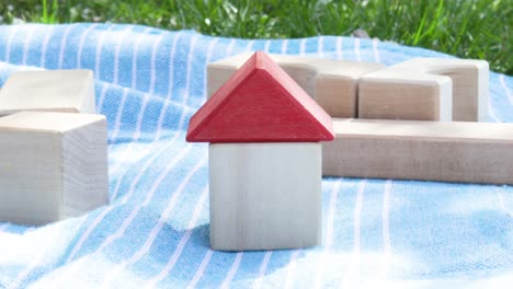 buy, rent, house insurance concept. a hand is putting red roof on a house from wooden eco cube toys. toys on blanket in park, nature background. sunny spring day