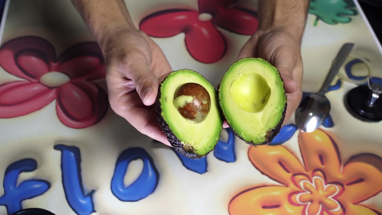 Primo piano dall'alto di mani maschili che tengono due metà di un avocado fresco e verde sopra un tavolo da cucina floreale e colorato. Preparazione di cibo vegetariano sano al chiuso con un cucchiaio di metallo visibile nelle vicinanze