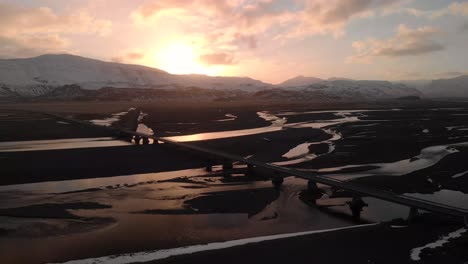 majestic aerial drone view of long straight bridge over several rivers in cold iceland. golden hour time after sunset, snow mountains and beutiful wild landscape