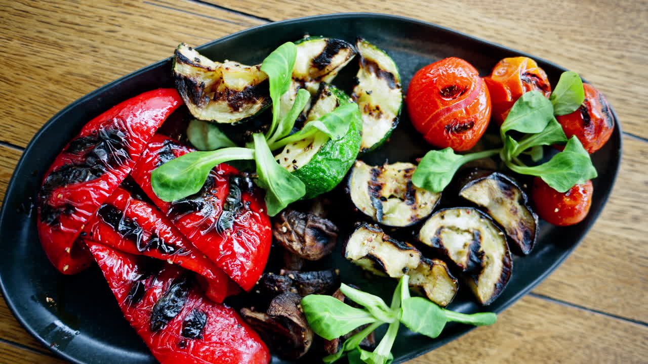 A black plate of grilled peppers, cherry tomatoes, zucchini, eggplant and mushrooms on a table