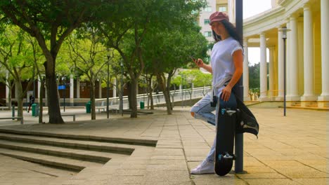 mujer joven usando teléfono móvil mientras se apoya en el poste con skateboard 4k