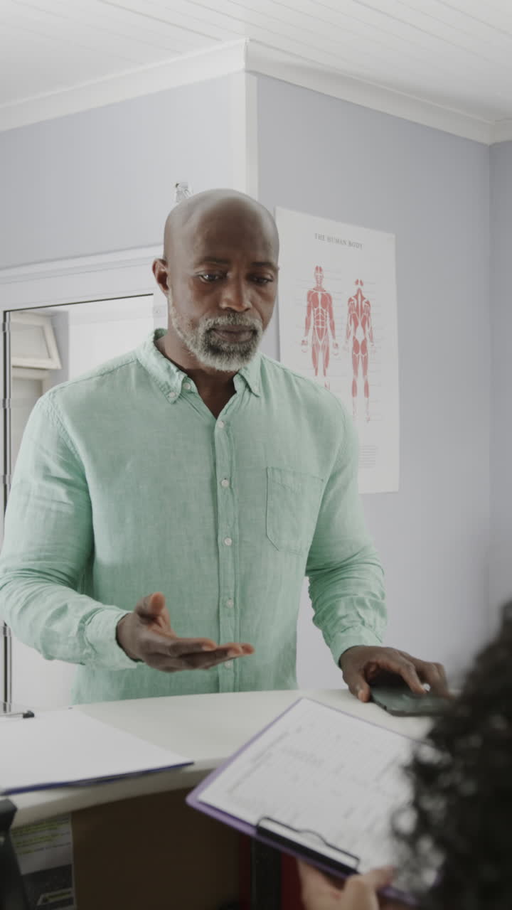 Vertical video of diverse medical receptionist and male patient talking at front desk, slow motion
