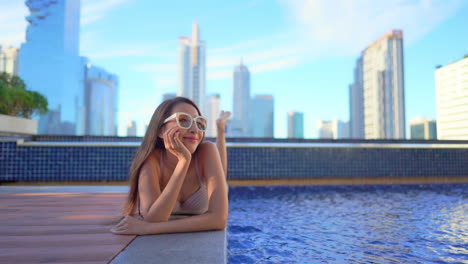 Mujer-Asiática-Exótica-Sexy-En-Traje-De-Baño-Disfrutando-De-La-Piscina-En-La-Azotea-Con-Edificios-Modernos-De-La-Ciudad-En-El-Fondo,-Cámara-Lenta-De-Marco-Completo