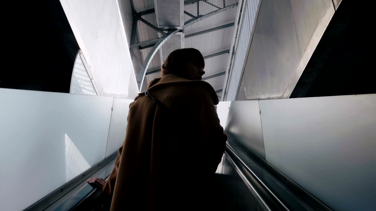 Low angle rear view young elegant tourist woman looks back riding modern escalator at a metro station slow motion.
