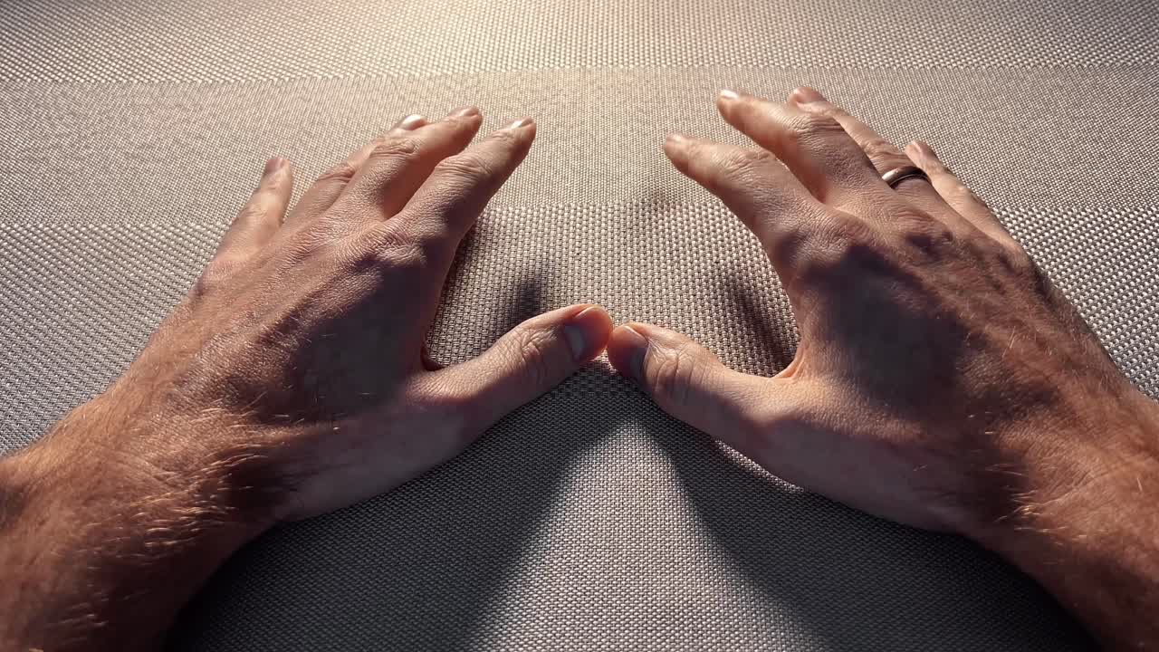 A cinematic close-up of a man's hands resting peacefully on a grey textured fabric. Strong sunlight creates deep shadows and highlights skin details, evoking a mood of contemplation and rest