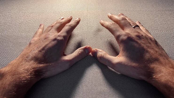A cinematic close-up of a man's hands resting peacefully on a grey textured fabric. Strong sunlight creates deep shadows and highlights skin details, evoking a mood of contemplation and rest