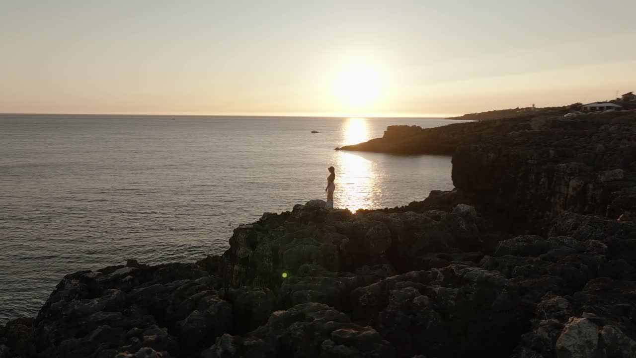 Aerial orbit of a woman in white standing motionless on coastal rocks during sunset, facing the horizon along Portugal’s coast