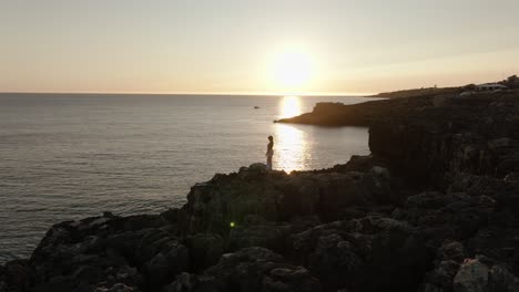 aerial orbit of a woman in white standing motionless on coastal rocks during sunset, facing the horizon along portugal’s coast