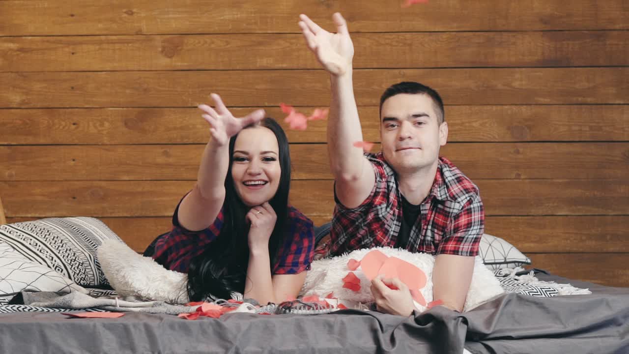 Joyful man and woman are laying on the bed and scattering paper hearts for Valentine's Day. Love story. Slow motion.