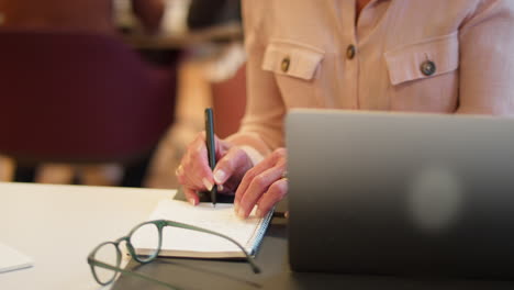 mujer de negocios madura trabajando en una computadora portátil en un escritorio en la oficina haciendo notas en un cuaderno