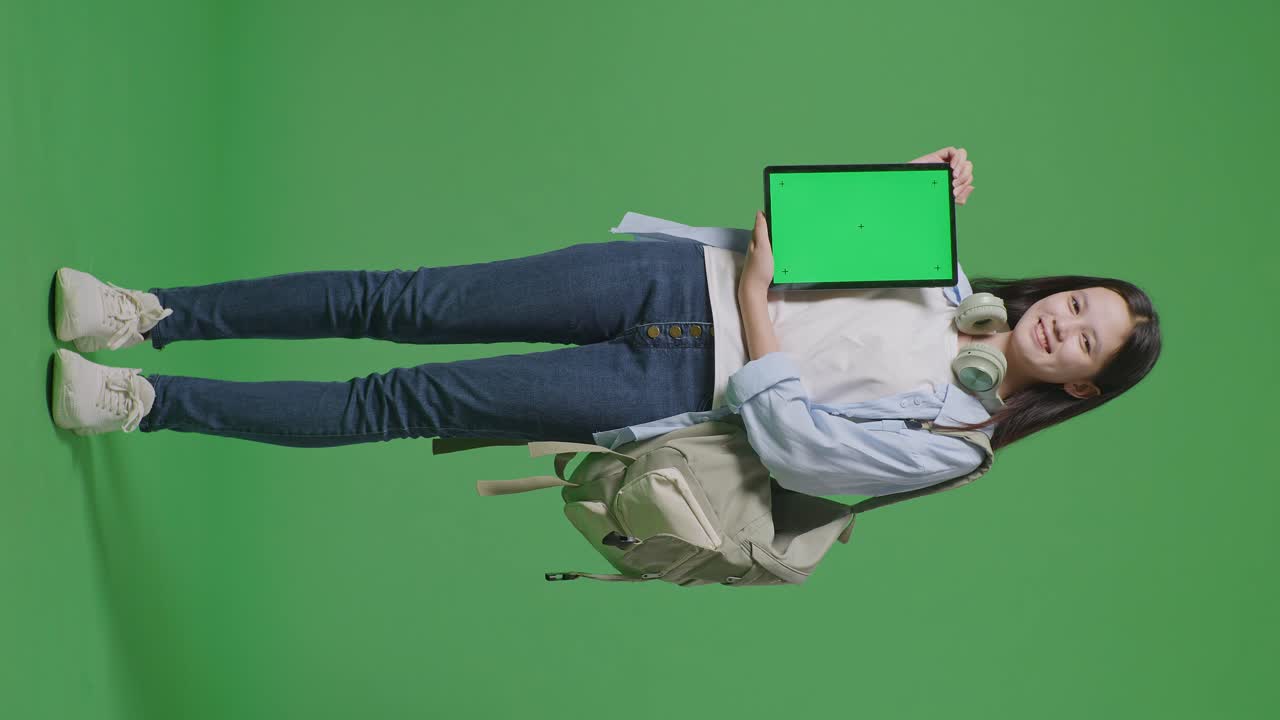 cuerpo lleno de estudiante adolescente asiática con una mochila sonriendo y mostrando la tableta de pantalla verde a la cámara mientras estaba de pie en el estudio de fondo de pantalla verde
