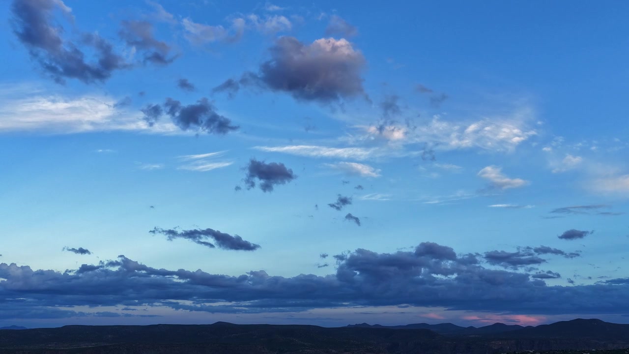 4K drone timelapse, der fanger det sidste aftenlys, som bølger hen over en blød, solbeskinnet sky. Mørke, lavtliggende skylag bevæger sig over et silhuetteret højtliggende ørkenbjerglandskab i skumringen