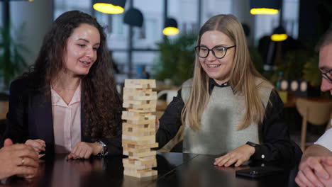 group of colleagues playing jenga during the lunch break. two women and man sitting at the table and playing board game in cafeteria.
