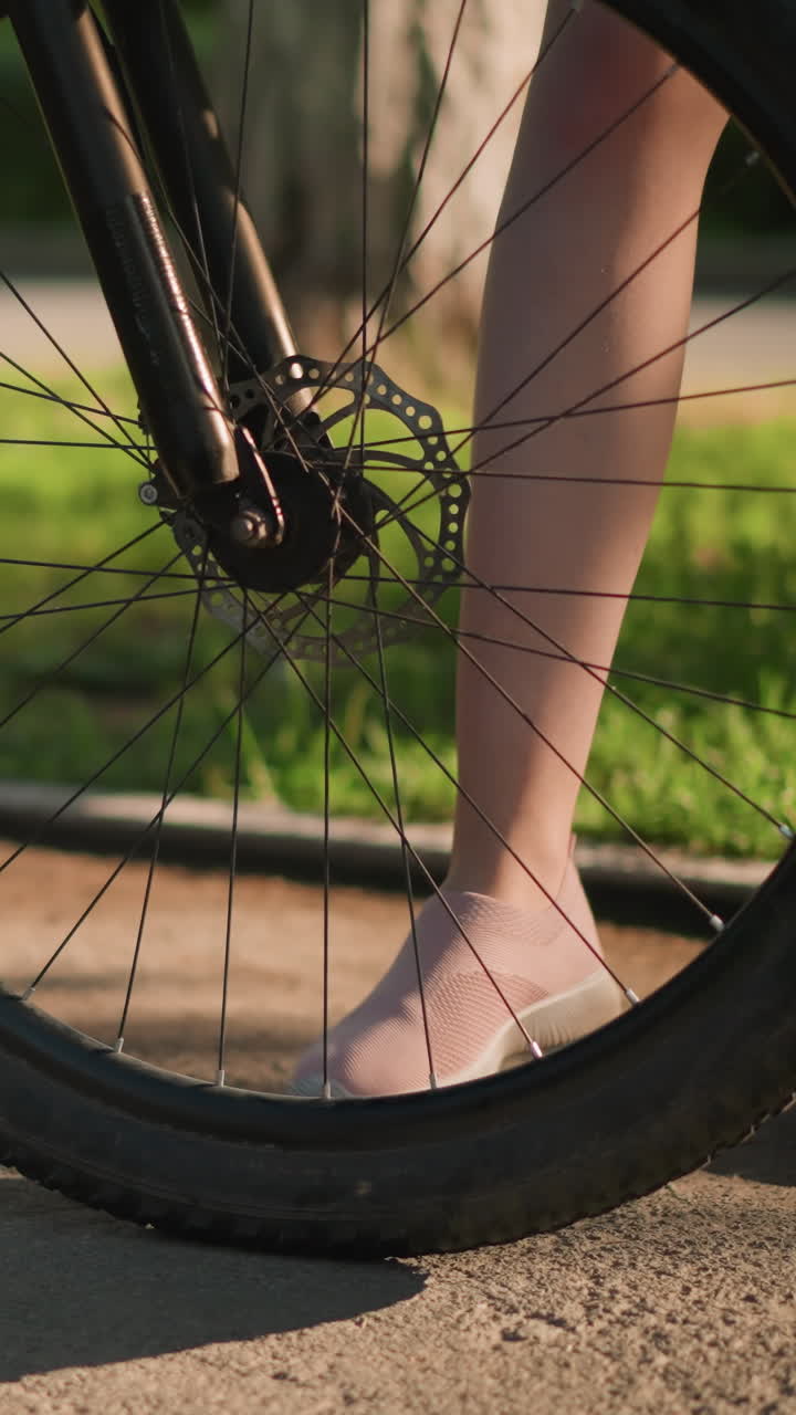 Close-up of someone's legs as they kick the bicycle tire to check air pressure, the background features lush greenery, adding a vibrant touch to the scene