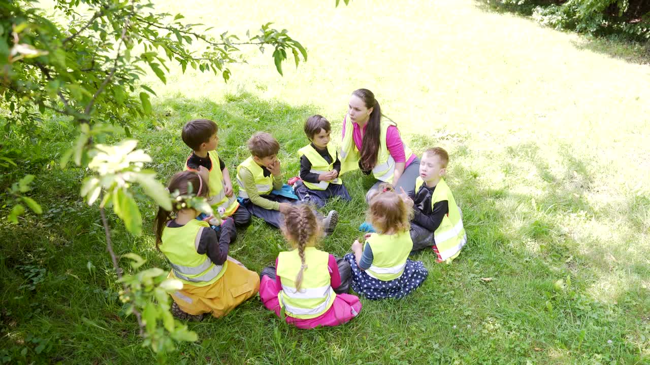 grupo de niños pequeños sentados en círculo con un tutor y jugando juegos juntos un césped en un parque o bosque. jardín de infantes una caminata o viaje. los niños se sientan en el prado de hierba se divierten.