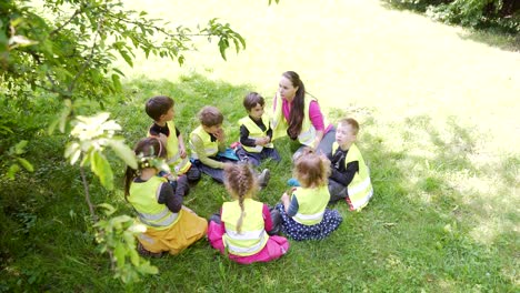 group little children sitting a circle with a tutor and playing games together a lawn in a park or forest. kindergarten a hike or trip. kids sit on the grass meadow have fun.