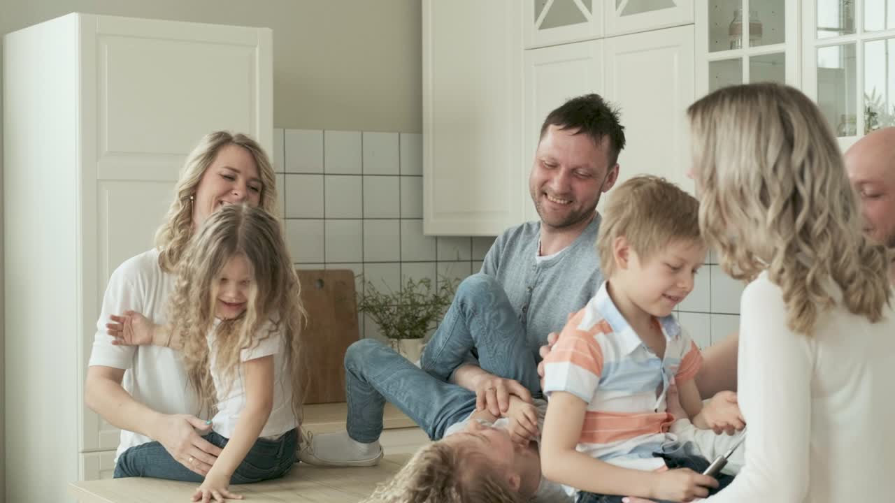 Big happy Caucasian family of three generations gathered together in kitchen at home, talking, laughing. Grandma and grandpa are playing with grandchildren. Parents and children spend time together