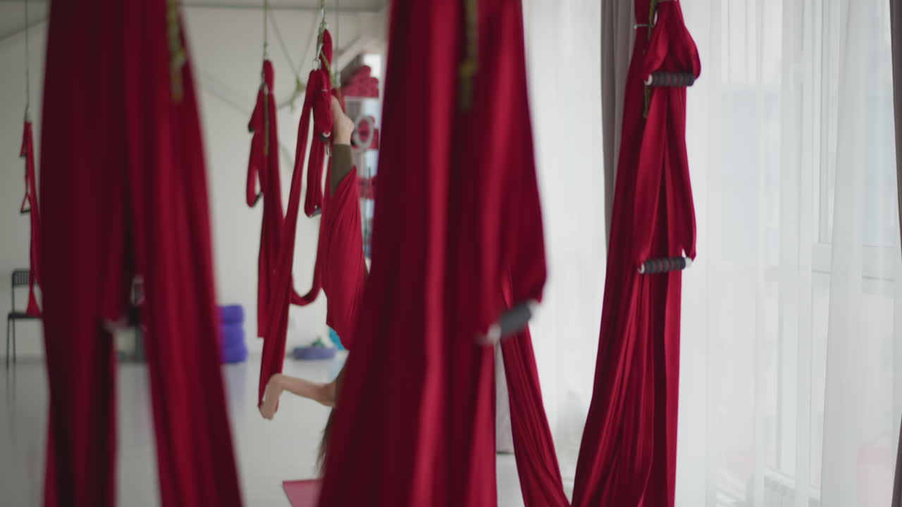 Flexible woman in sportswear stretches inverted in red aerial silk hammocks suspended from ceiling by sheer curtains and windows demonstrating core strength balance in modern wellness studio