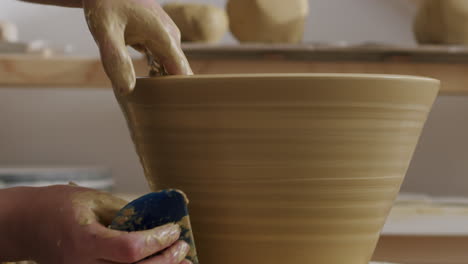 working with a hand scraper on a big brown clay vase, pulling the scraper from the bottom to the top to get rid of material